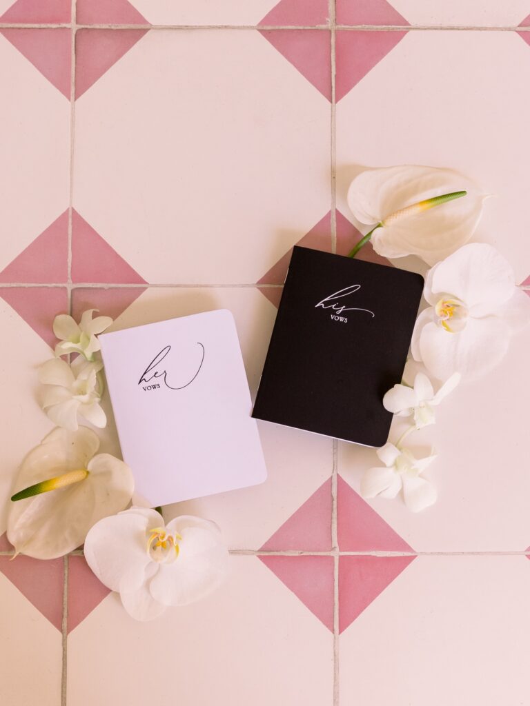 His and her vow books on a pink diamond-shaped tile floor with loose flowers